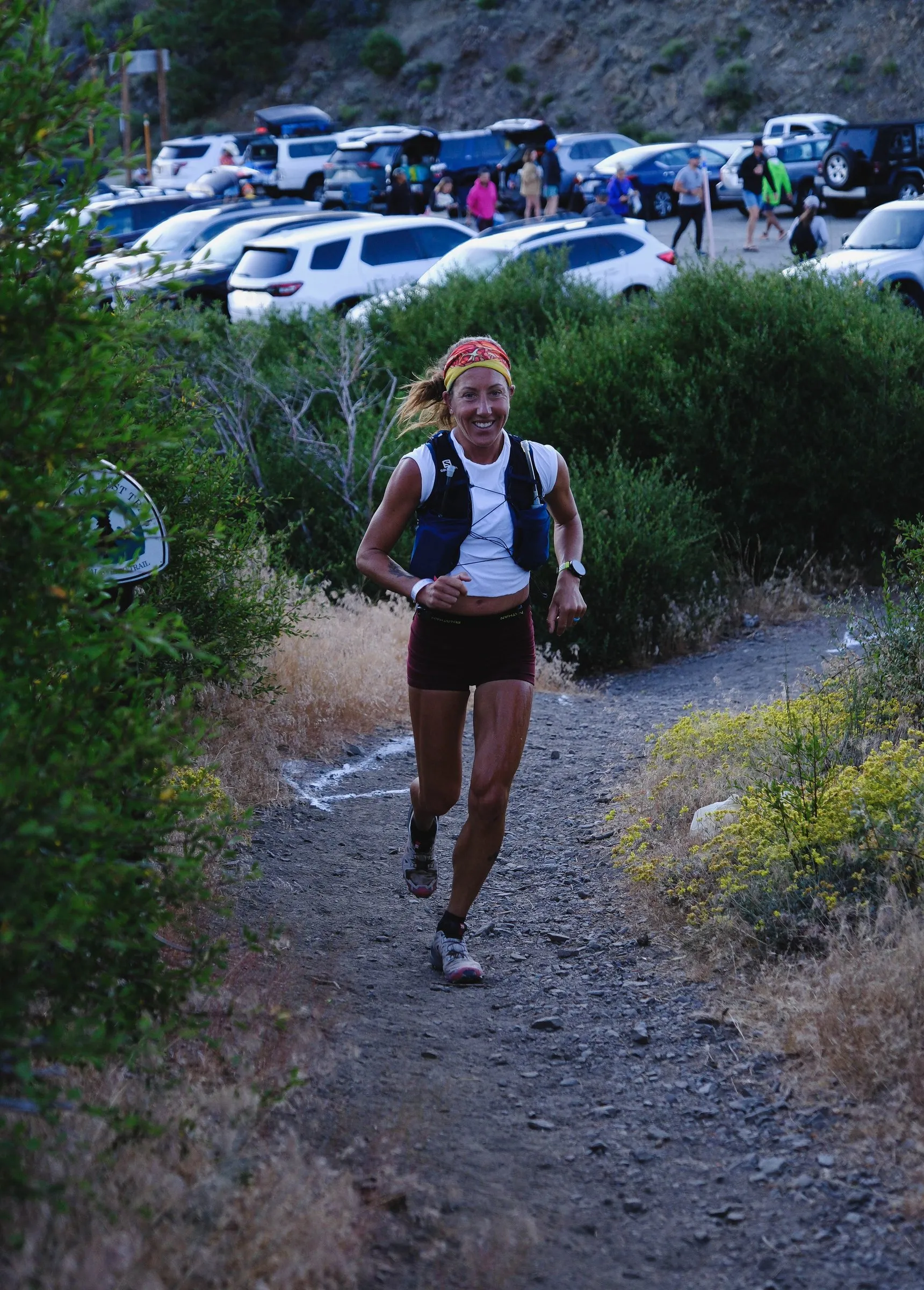 Megan Eckert running up a trail from a crowded aid station in race vest and headband